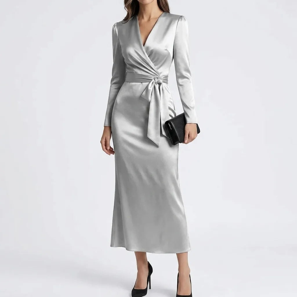 A woman models a long-sleeve wrap midi dress in a light silver-gray satin fabric, styled with a black clutch and black heels against a plain light gray studio background.