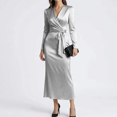 A woman models a long-sleeve wrap midi dress in a light silver-gray satin fabric, styled with a black clutch and black heels against a plain light gray studio background.