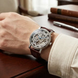 A wrist wearing a skeleton-style analog watch with a brown leather strap and silver-toned case, resting on a wooden desk with books and a pen in the background.