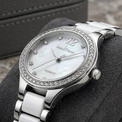 A silver-tone quartz watch with a white mother-of-pearl dial, a crystal-studded bezel, and a matching silver link bracelet, displayed on a dark textured cushion.
