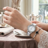 A silver-tone stainless steel watch with a white dial and crystal-embellished bezel, worn on a woman's wrist alongside a black clover charm bracelet, set in a softly lit cafe environment.