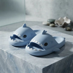 A pair of light blue shark-shaped slide slippers with white teeth details, displayed on a gray marble block with seashells in the background.