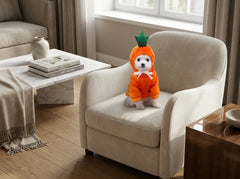 A small white dog wearing an orange carrot costume sits on a beige armchair in a modern living room setting.