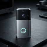 A modern wireless video doorbell with a silver and black finish, standing on a dark desk. It features a glowing blue circular light on the front, a central camera lens, and a small speaker grille.