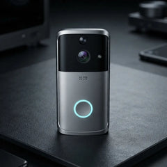 A modern wireless video doorbell with a silver and black finish, standing on a dark desk. It features a glowing blue circular light on the front, a central camera lens, and a small speaker grille.