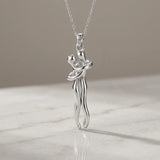 A silver pendant necklace featuring a stylized dancing couple, suspended from a delicate silver chain and resting on a light marble surface against a neutral beige background.