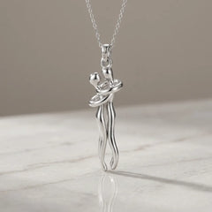 A silver pendant necklace featuring a stylized dancing couple, suspended from a delicate silver chain and resting on a light marble surface against a neutral beige background.