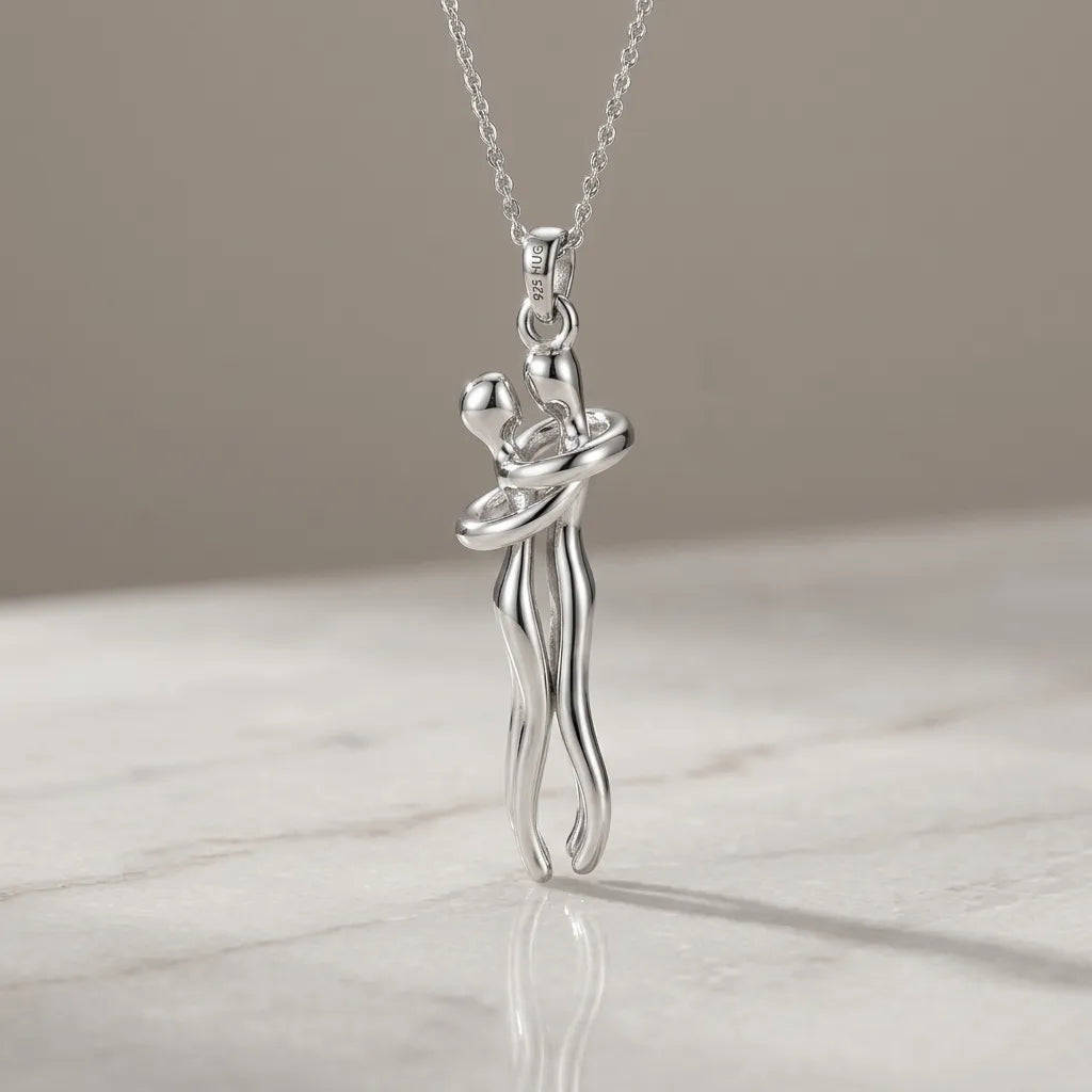 A silver pendant necklace featuring a stylized dancing couple, suspended from a delicate silver chain and resting on a light marble surface against a neutral beige background.