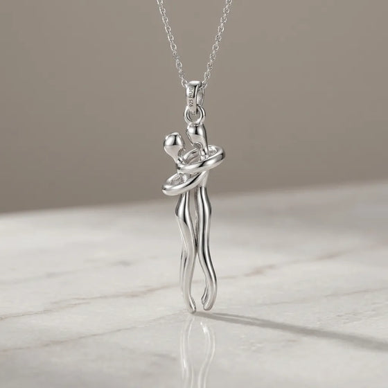 A silver pendant necklace featuring a stylized dancing couple, suspended from a delicate silver chain and resting on a light marble surface against a neutral beige background.