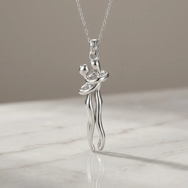 A silver pendant necklace featuring a stylized dancing couple, suspended from a delicate silver chain and resting on a light marble surface against a neutral beige background.