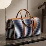 A navy and white horizontally striped duffel bag with tan leather handles and trim, resting on a dark marble surface against a neutral indoor background.
