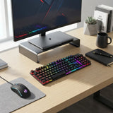 A black mechanical keyboard with vibrant rainbow RGB backlighting sits on a light wood desk next to a matching gaming mouse on a gray mousepad. The desk setup is styled with a monitor on a silver riser, a notebook, and a small succulent.