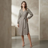 A woman models a long-sleeve wrap dress with a small-scale geometric pattern in gray and beige, standing in a minimalist studio with textured beige walls.