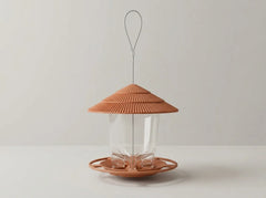 A modern bird feeder with a textured terracotta-colored lid and base, a clear glass body, and a thin metal hanging loop, set against a neutral light gray background.