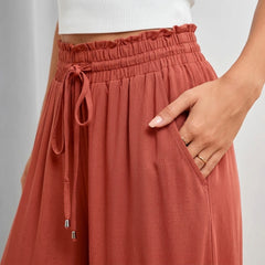 A close-up of a person wearing terracotta-colored wide-leg pants with a ruffled elastic waistband, drawstring, and side pockets. The person is also wearing a white ribbed crop top, and their hand is resting in the pocket.