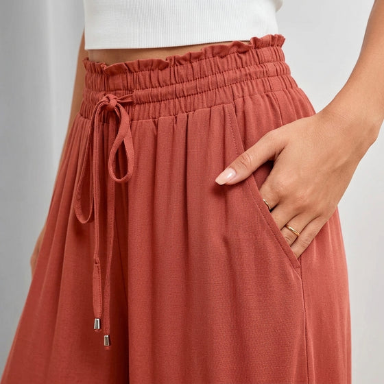 A close-up of a person wearing terracotta-colored wide-leg pants with a ruffled elastic waistband, drawstring, and side pockets. The person is also wearing a white ribbed crop top, and their hand is resting in the pocket.