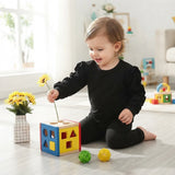 A toddler wearing a black long-sleeve top and black pants sits on a light wood floor, holding a yellow flower above a colorful shape-sorting cube toy. The room is brightly lit with natural light from a window, and other toys and a vase of yellow flowers are visible in the background.