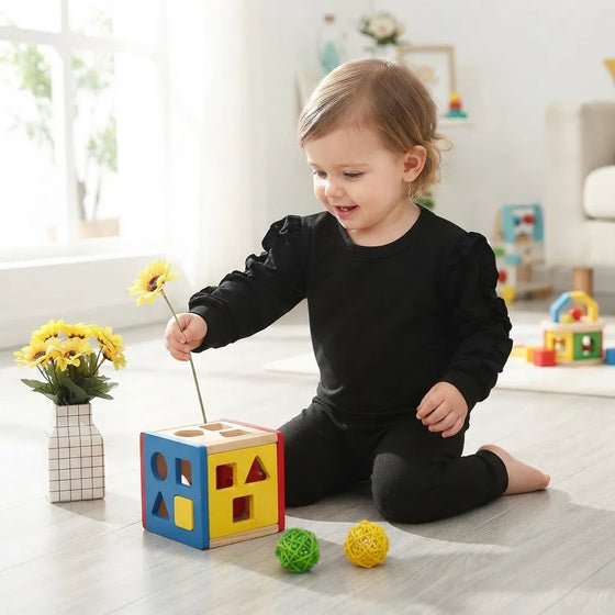 A toddler wearing a black long-sleeve top and black pants sits on a light wood floor, holding a yellow flower above a colorful shape-sorting cube toy. The room is brightly lit with natural light from a window, and other toys and a vase of yellow flowers are visible in the background.