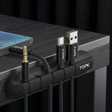 A black TOPK cable organizer clip mounted on a dark wood desk, holding a 3.5mm audio jack, a USB-C cable, and a USB-A cable.