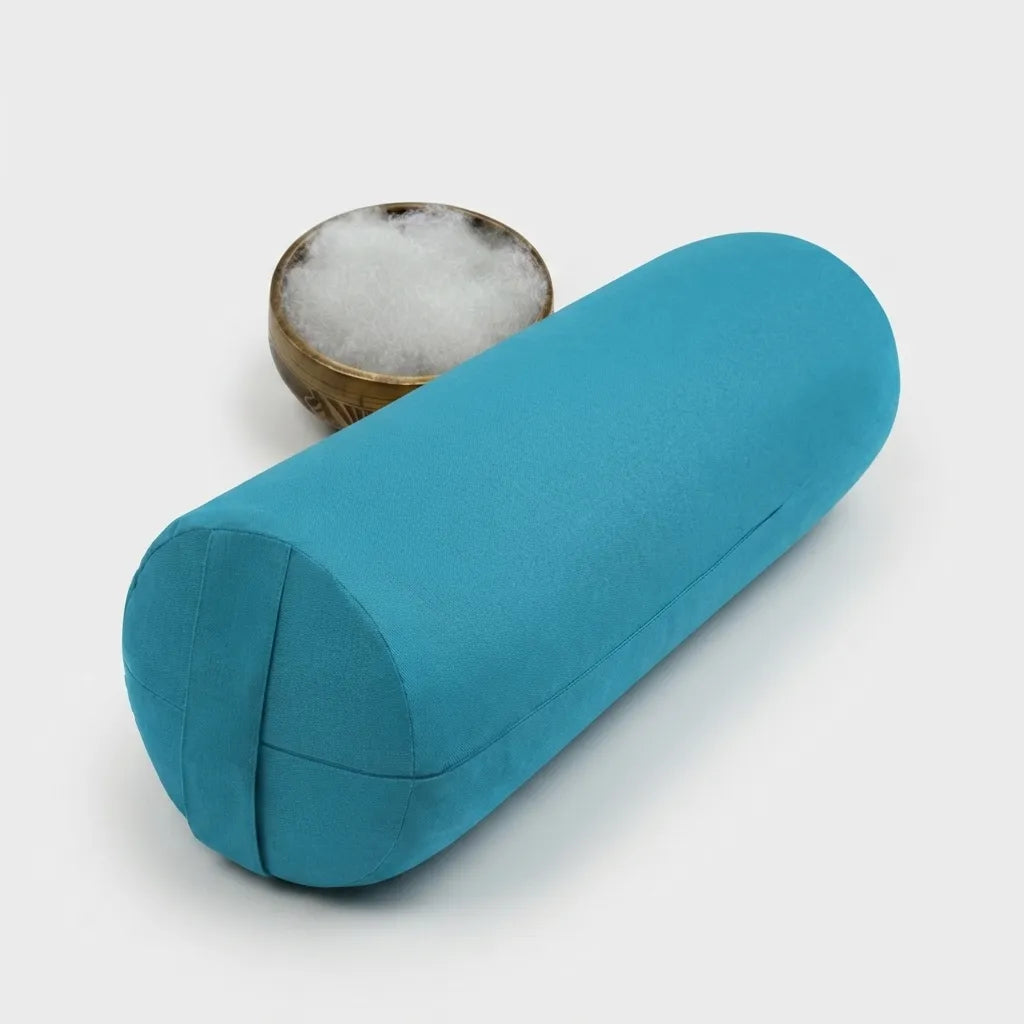 A teal blue cylindrical yoga bolster pillow shown on a light gray background, with a small wooden bowl of white cotton padding behind it.