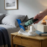 A white wireless charging dock sits on a wooden bedside table, charging a white earbud case and a smartphone. A hand holds a blue smartphone above the dock, demonstrating wireless charging.