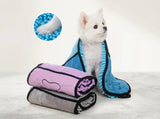 A small white dog sits wrapped in a blue microfiber towel with black trim, while two rolled towels in pink and gray are stacked in front. A circular inset shows a close-up of blue microfiber texture.
