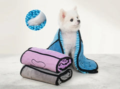 A small white dog sits wrapped in a blue microfiber towel with black trim, while two rolled towels in pink and gray are stacked in front. A circular inset shows a close-up of blue microfiber texture.