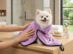 A small white dog sits on a wooden bathroom counter while being dried with a lavender microfiber towel with black trim. A person's hands are visible adjusting the towel.