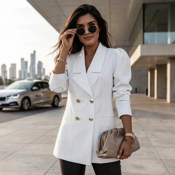 A woman wearing a white double-breasted blazer with gold buttons, black pants, and sunglasses, holding a beige clutch bag in an outdoor urban setting.