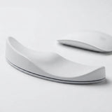 A white ergonomic wrist rest with a curved, wave-like design, shown on a clean white surface with a blurred white computer mouse in the background.