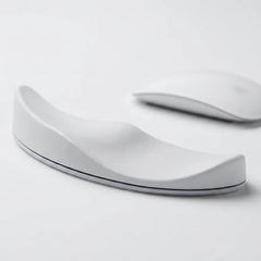 A white ergonomic wrist rest with a curved, wave-like design, shown on a clean white surface with a blurred white computer mouse in the background.