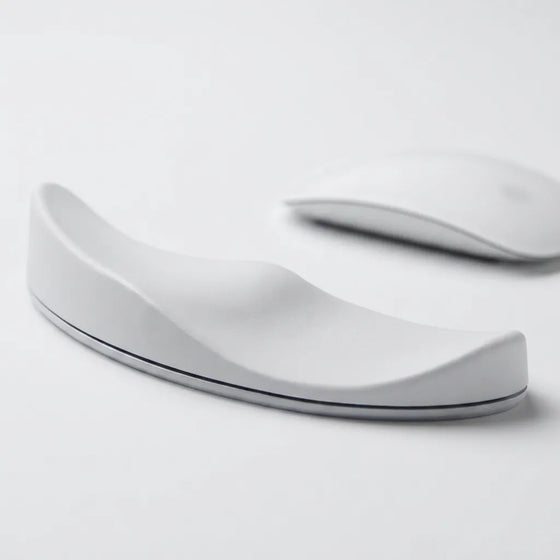 A white ergonomic wrist rest with a curved, wave-like design, shown on a clean white surface with a blurred white computer mouse in the background.