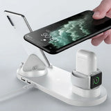 A white multi-device wireless charging stand with a phone charging dock, a watch charging cradle, and a power cable port, shown with a smartphone and a white earbud case on a light gray background.