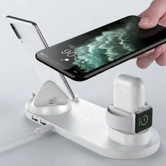 A white multi-device wireless charging stand with a phone charging dock, a watch charging cradle, and a power cable port, shown with a smartphone and a white earbud case on a light gray background.