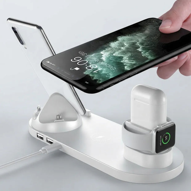 A white multi-device wireless charging stand with a phone charging dock, a watch charging cradle, and a power cable port, shown with a smartphone and a white earbud case on a light gray background.