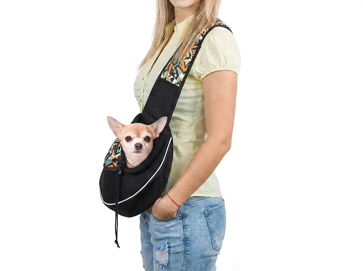 A woman wearing a light yellow shirt and blue jeans models a black pet sling carrier with a small Chihuahua dog peeking out from the front. The carrier has a wide black body with a colorful patterned strap and white piping details.