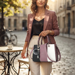 A woman in a dusty rose blazer and beige trousers carries a structured handbag with a color-blocked design in black, dusty purple, and white, featuring a black faux-croc texture and a fluffy pom-pom charm.