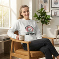 A woman sitting in a wooden armchair in a bright living room, wearing a white crewneck sweatshirt with a graphic that says 'EVERYTHING IS POSSIBLE WITH A CAT BEST FRIEND' around a pink cat illustration. She is holding a beige mug.