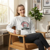 A woman sitting in a wooden armchair in a bright living room, wearing a white crewneck sweatshirt with a graphic that says 'EVERYTHING IS POSSIBLE WITH A CAT BEST FRIEND' around a pink cat illustration. She is holding a beige mug.