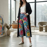 A woman models a pair of wide-leg cropped jeans featuring a vibrant multicolor floral print on a blue denim base, styled with a white top, a black open-front cardigan, and white slip-on shoes in a modern, sunlit room.