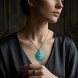 A close-up lifestyle shot of a woman wearing a silver crescent moon pendant necklace with a glowing turquoise light inside, set against a dark, moody background.