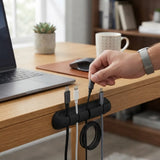 A black cable management holder mounted on the edge of a wooden desk, holding several cables. A person's hand is shown plugging a cable into the holder.